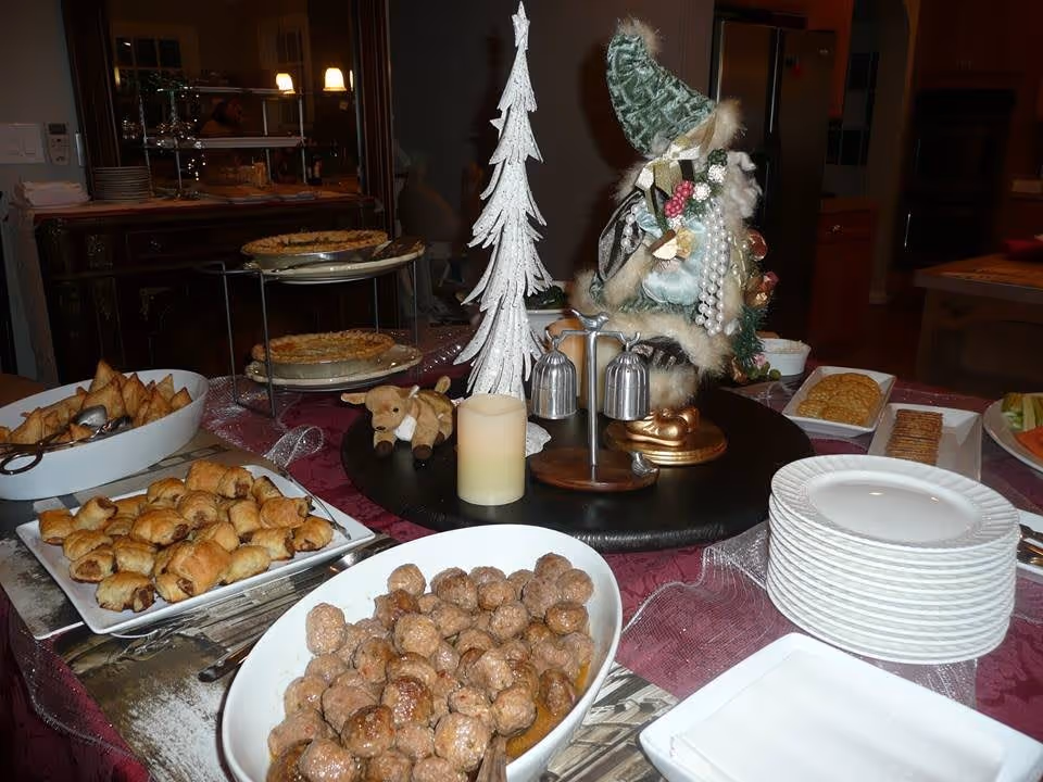 A buffet table set with various foods including meatballs, pastries, crackers, and pies. There are stacks of white plates and a decorative centerpiece featuring a white Christmas tree, a small stuffed reindeer, a candle, and a figurine dressed in green with pearls. The setting appears to be indoors with kitchen cabinets and appliances visible in the background.
