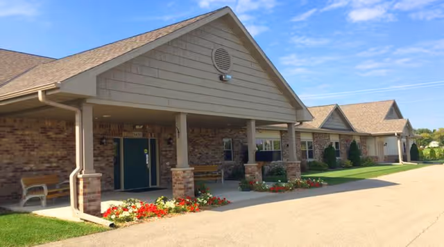 Exterior view of a single-story senior living facility building with a covered entrance supported by columns, brick and siding walls, flower beds with red and white flowers, and a clear blue sky.
