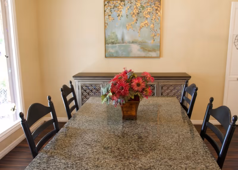 A dining room with a granite table surrounded by six black wooden chairs. A vase with pink flowers is placed in the center of the table. Behind the table is a decorative sideboard with intricate panel designs, and above it hangs a painting of a serene landscape with trees and a path.