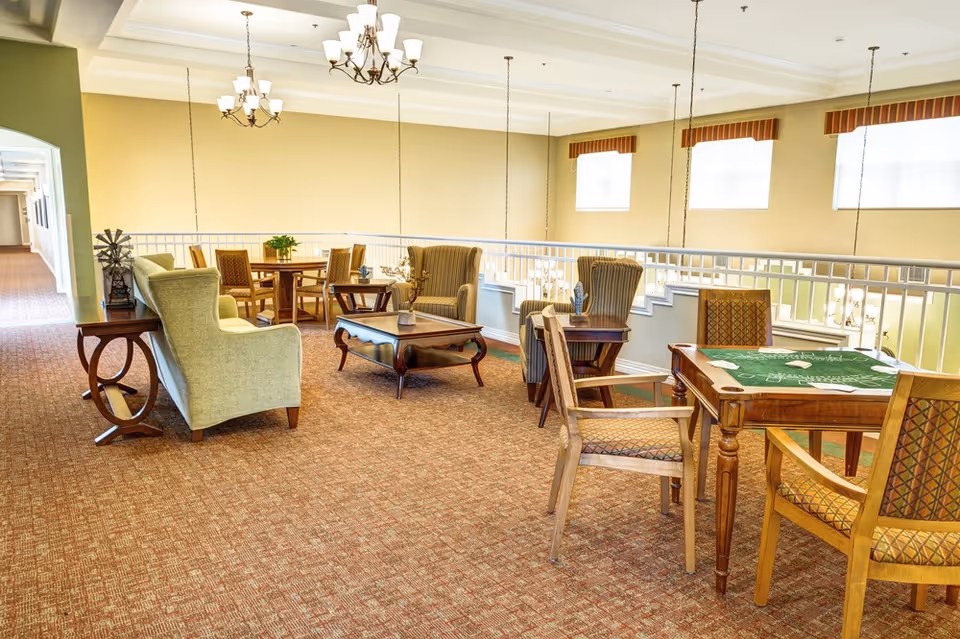 Bright communal lounge with sofas, armchairs, and tables arranged for dining and games in a senior living facility.