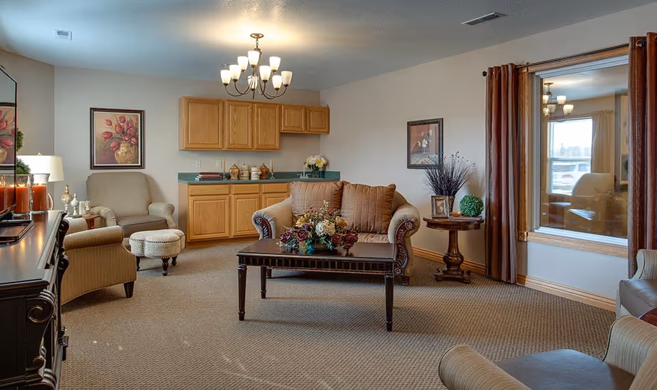 A cozy senior living facility common area with beige walls and carpet. The room features a comfortable loveseat with two brown cushions, two armchairs, a small ottoman, a wooden coffee table with a floral centerpiece, and a side table with decorative items. There are framed floral paintings on the walls, a chandelier hanging from the ceiling, and a large window with brown curtains reflecting the room.