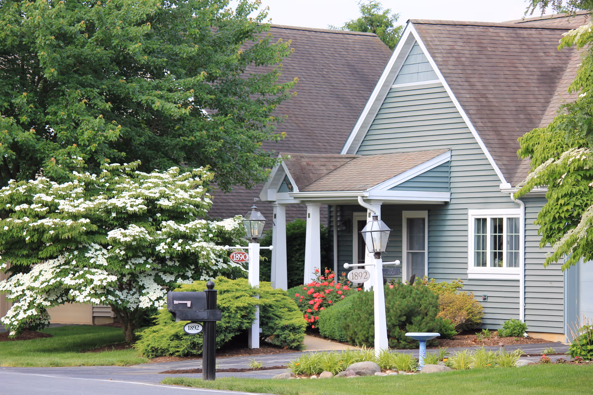 Exterior view of a residential building with gray siding and a brown roof, surrounded by green trees and bushes with white flowers. There are two black mailboxes and two white posts with lanterns and house number signs (1890 and 1892) near the sidewalk.