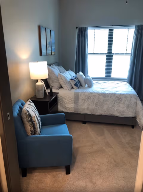 A cozy bedroom with a neatly made bed featuring multiple pillows and a floral-patterned bedspread. Next to the bed is a dark wooden nightstand with a white table lamp. A blue upholstered armchair with a decorative pillow is positioned near the nightstand. A large window with closed blinds and blue curtains allows natural light into the room. Two pieces of wall art hang above the bed.