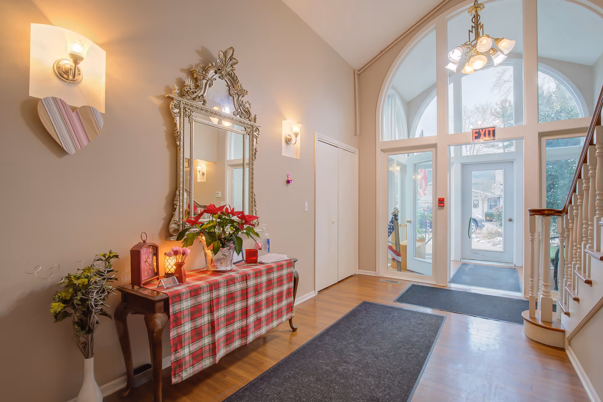 Interior entryway of a facility with a wooden floor, a decorative table covered with a red plaid cloth, a large ornate mirror above the table, wall sconces with lights, a heart-shaped wall decoration, and a glass door with an exit sign above it. There is a staircase with white railings on the right side and mats on the floor.