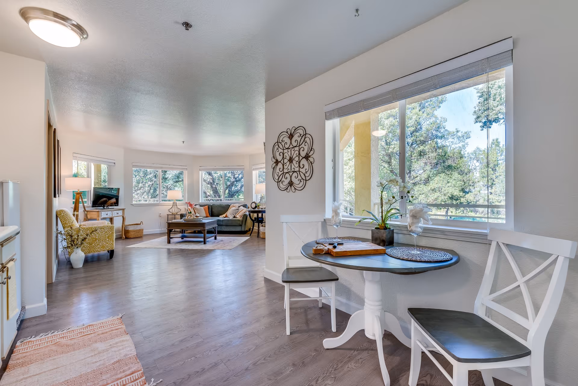 Bright and spacious senior living area with a small round dining table and two chairs near a large window overlooking trees. The room features wood flooring, a cozy seating area with a sofa, armchair, coffee table, and multiple windows allowing natural light to fill the space.
