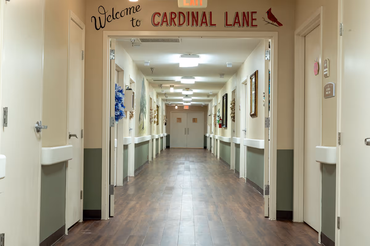 A long, well-lit hallway in a senior living facility with multiple doors on both sides. The walls are painted beige with a green lower half, and the floor is wood-patterned. Above the entrance to the hallway is a sign that reads 'Welcome to CARDINAL LANE' with a small cardinal bird graphic. There are handrails along the walls and some decorations hanging on the doors and walls.