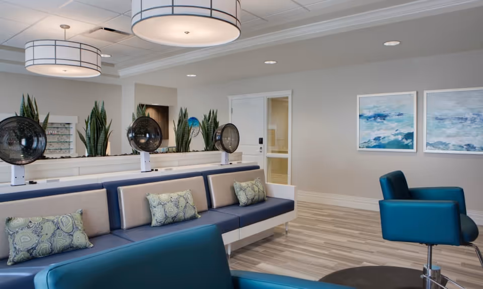 Interior view of a modern salon area in an assisted living facility featuring blue and beige cushioned seating with patterned pillows, three vintage-style hair dryers, potted plants, two abstract ocean-themed paintings on the wall, and large round ceiling lights.