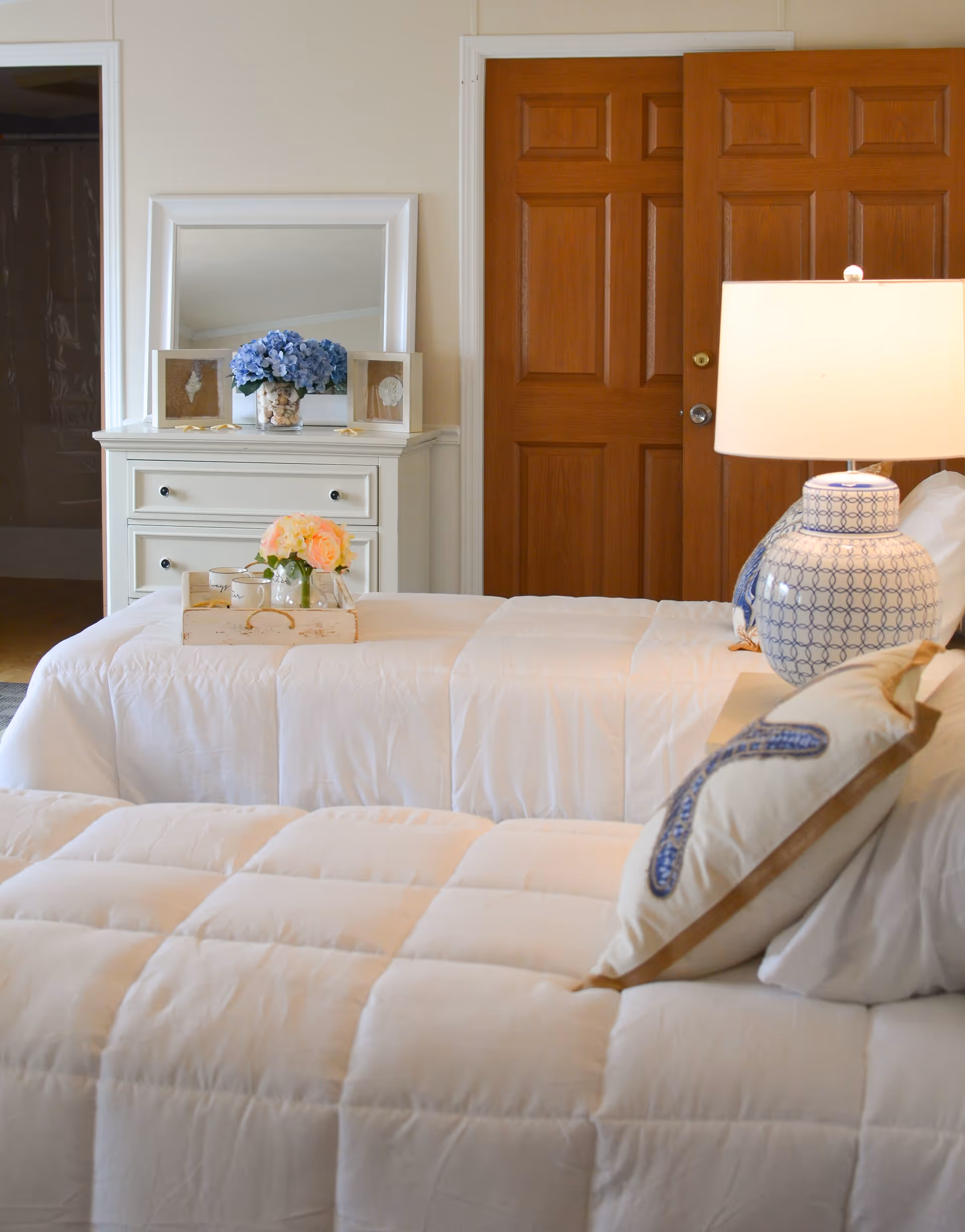 A cozy bedroom with two twin beds covered in white comforters. One bed has a decorative pillow and a blue and white patterned lamp on the nightstand. A white dresser with a mirror, blue flowers, and framed seashell pictures is against the wall. There are wooden closet doors in the background.