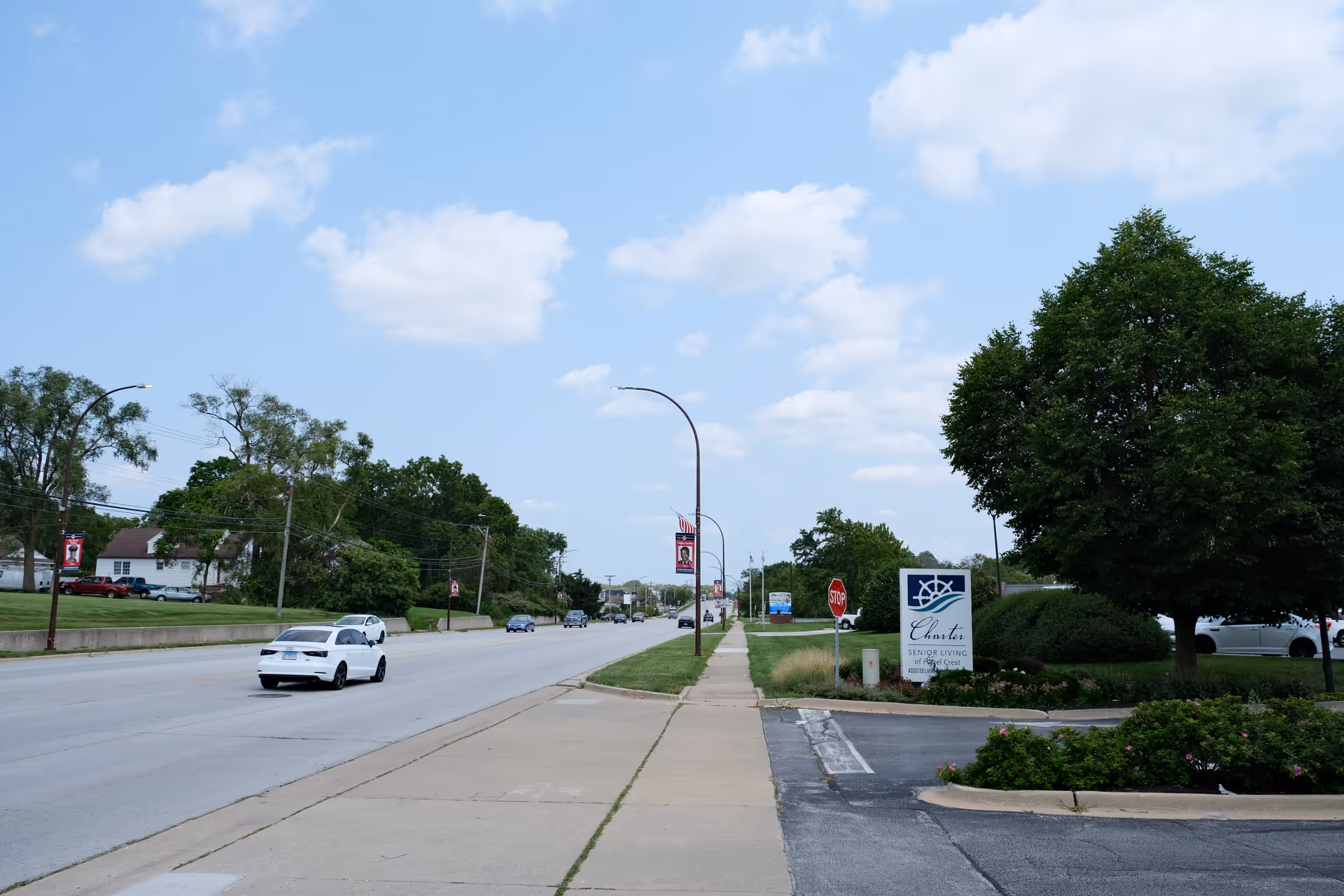 Street view showing a sidewalk and road with several cars driving. On the right side, there is a sign for Charter Senior Living of Hazel Crest surrounded by greenery and trees under a partly cloudy sky.