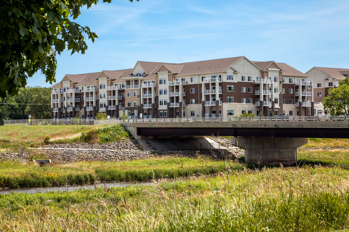 A large multi-story residential building with balconies, situated behind a bridge over a small river or creek, surrounded by grassy and wooded areas under a clear blue sky.