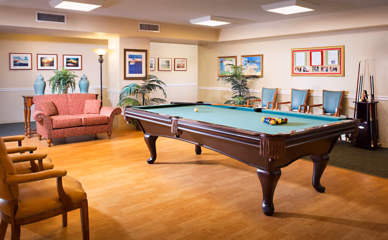 A well-lit recreational room featuring a pool table with balls arranged for a game. The room has wooden flooring, several chairs including a red patterned loveseat, and framed pictures on the walls. There are also potted plants and a floor lamp adding to the cozy atmosphere.