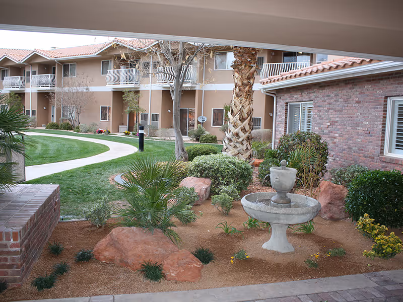Courtyard with a small stone fountain, landscaped rocks and plants in front of a two-story assisted living building with balconies.