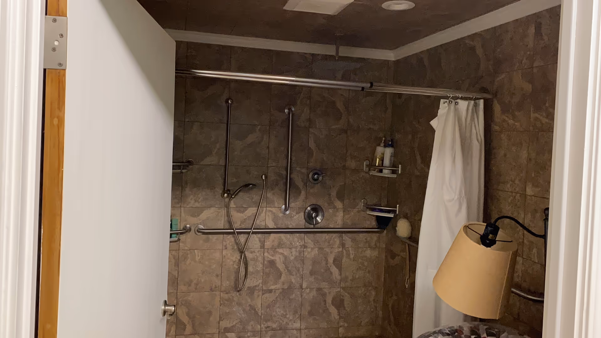 Accessible tiled shower with multiple grab bars, a handheld showerhead and a white curtain seen through a partially open door.