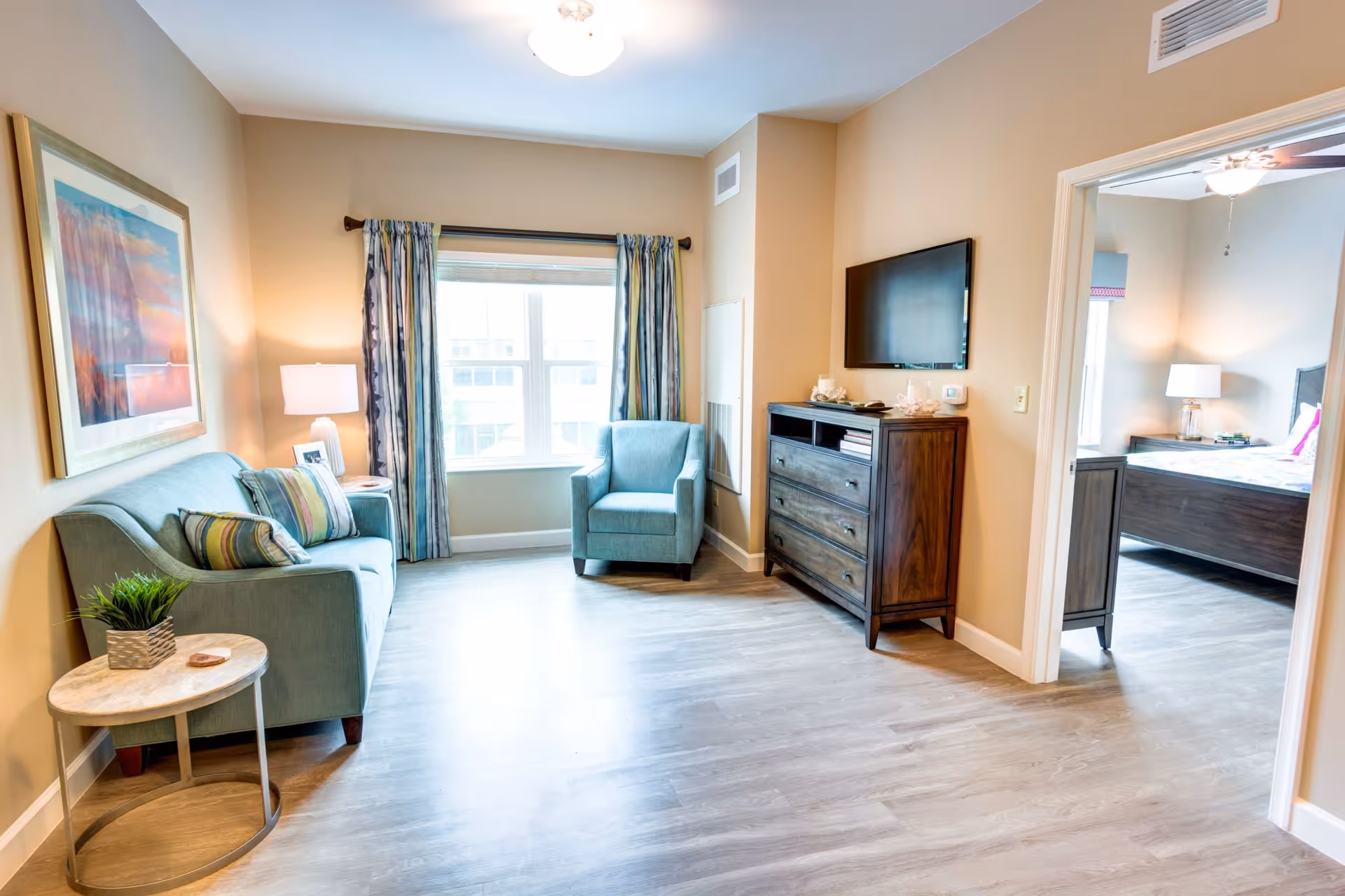 A bright living room with light wood flooring, a teal sofa with striped pillows, a matching armchair, a small round side table with a plant, a framed artwork on the wall, and a window with blue and white curtains. A wooden dresser with a flat-screen TV mounted above it is against the opposite wall. An open doorway leads to a bedroom with a bed, nightstand, and lamp.