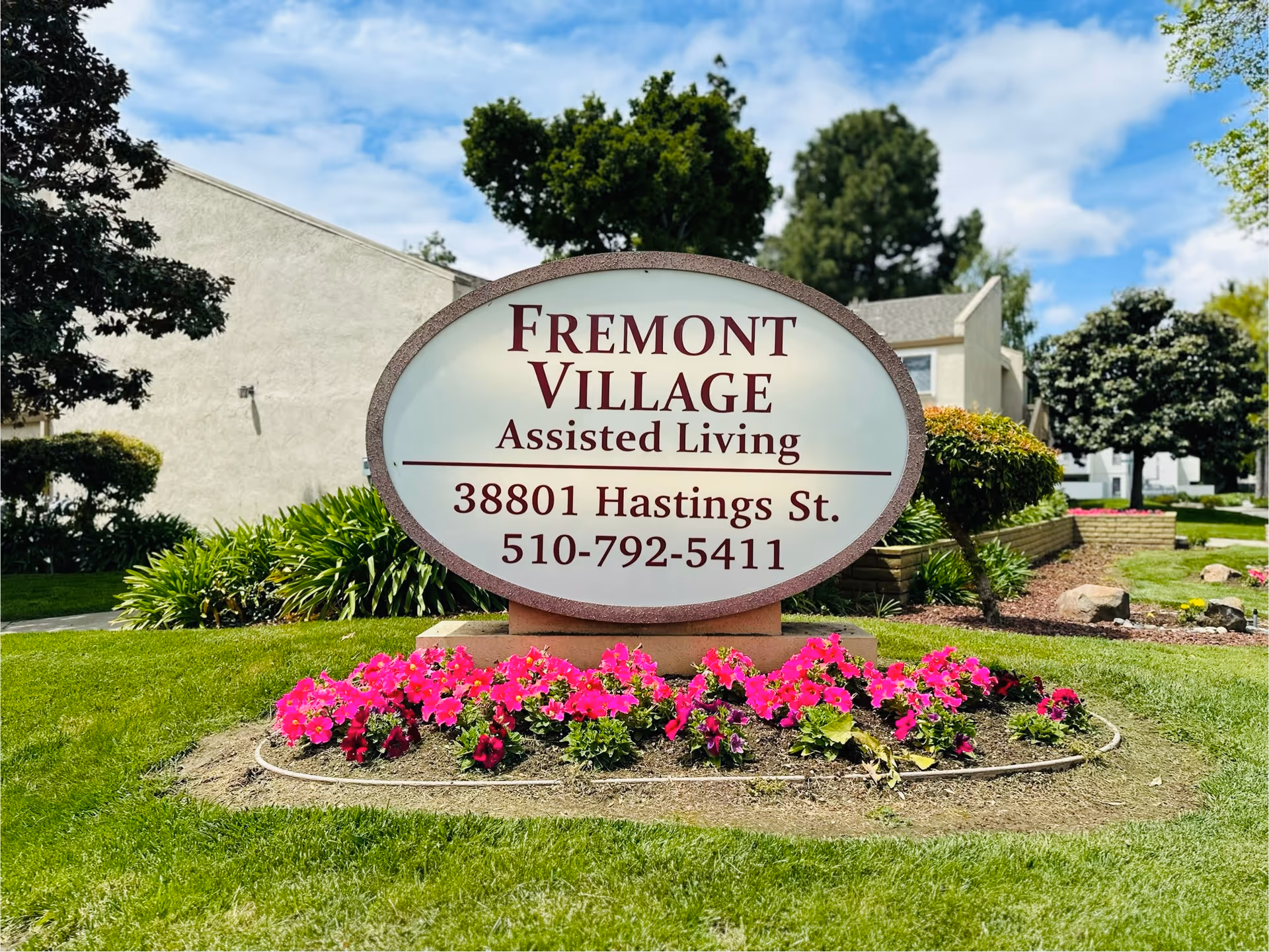 Oval Fremont Village Assisted Living sign showing address and phone number placed in a landscaped lawn with flowers and buildings behind it.