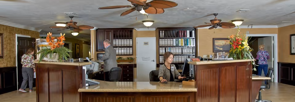 Reception area with a wooden front desk, a receptionist at a computer, other people, filing shelves and decorative plants.