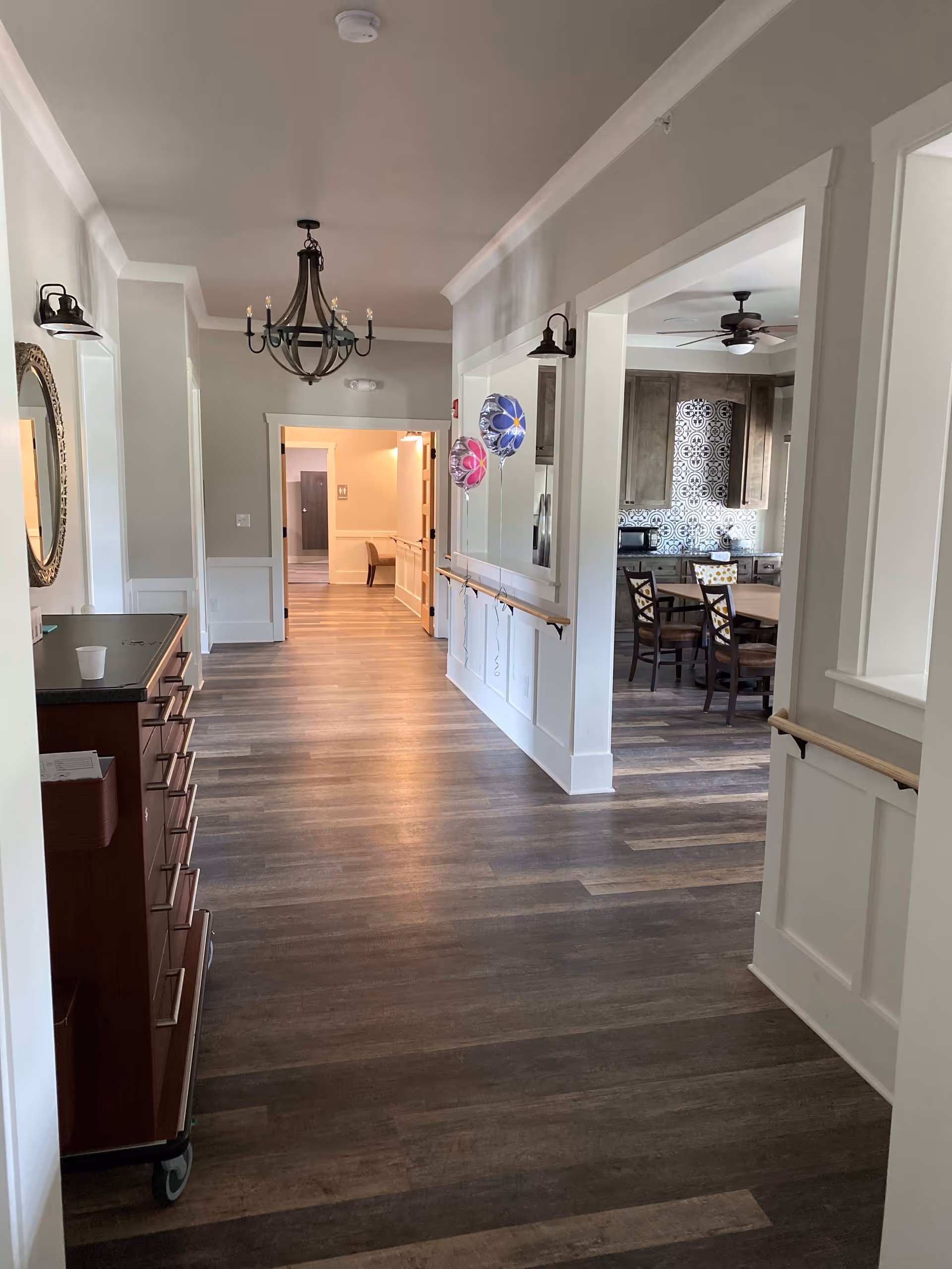 Interior hallway of a senior living facility with wooden flooring, white walls, and a chandelier hanging from the ceiling. On the right side, there is an open view into a kitchen area with a dining table and chairs, a ceiling fan, and patterned backsplash tiles. Two colorful balloons are attached to the wall along the hallway.