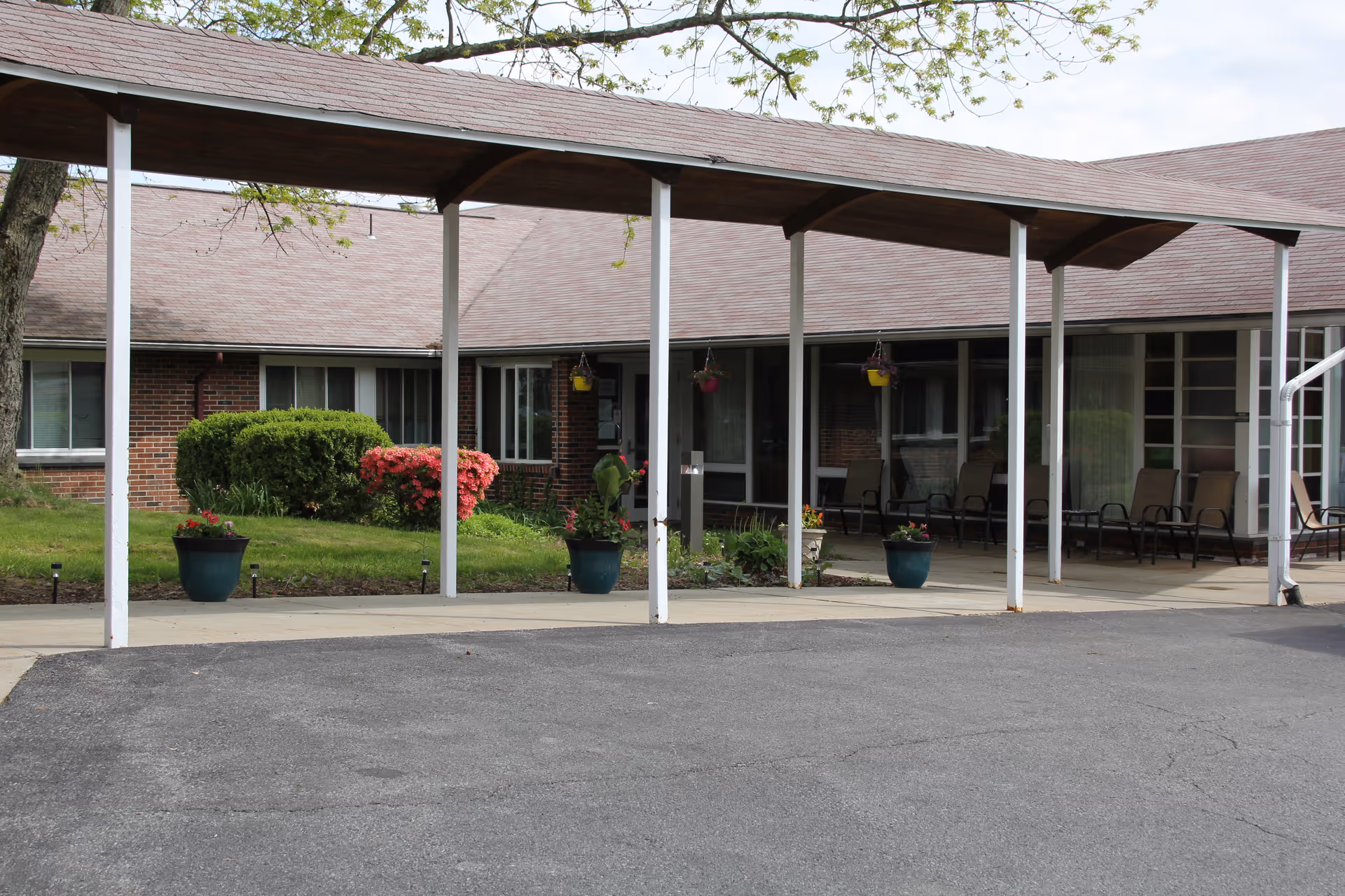 Exterior view of a single-story brick building with a covered walkway supported by white pillars. There are several potted plants and hanging flower baskets along the walkway, with bushes and flowering shrubs in the background. Several chairs are arranged under the covered area near the building entrance.