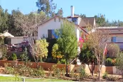 Two-story residential building with a landscaped front yard, trees, and a deck under a clear blue sky.