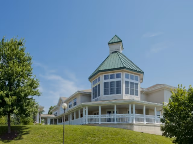 Exterior view of a senior living facility building with a green metal roof and large windows on a sunny day, surrounded by green grass and trees.