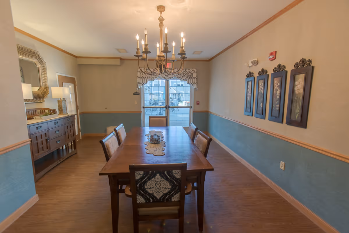 A dining room with a wooden table and six chairs, a chandelier hanging above the table, a sideboard with a lamp and mirror on the left wall, floral artwork on the right wall, and glass doors leading outside.