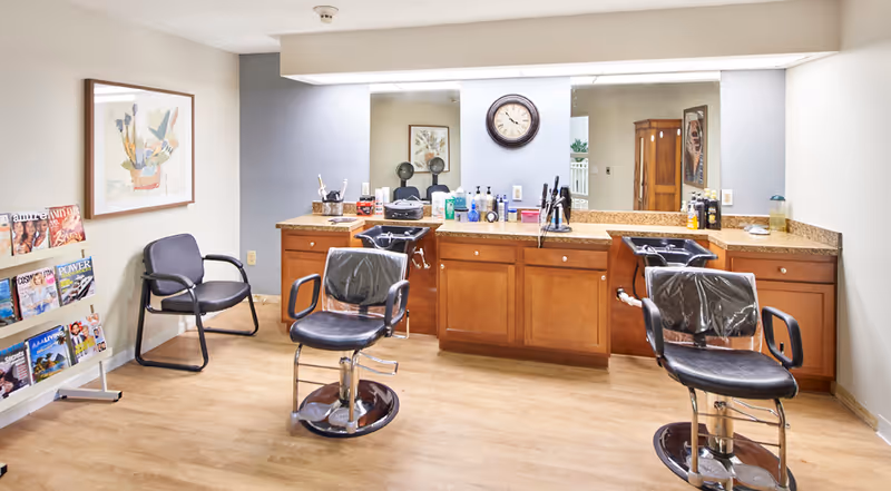 Interior view of a salon area in a senior living facility with two black salon chairs covered in plastic, a black waiting chair, a magazine rack with various magazines, a large mirror above a wooden counter with sinks and hair care products, and a wall clock.