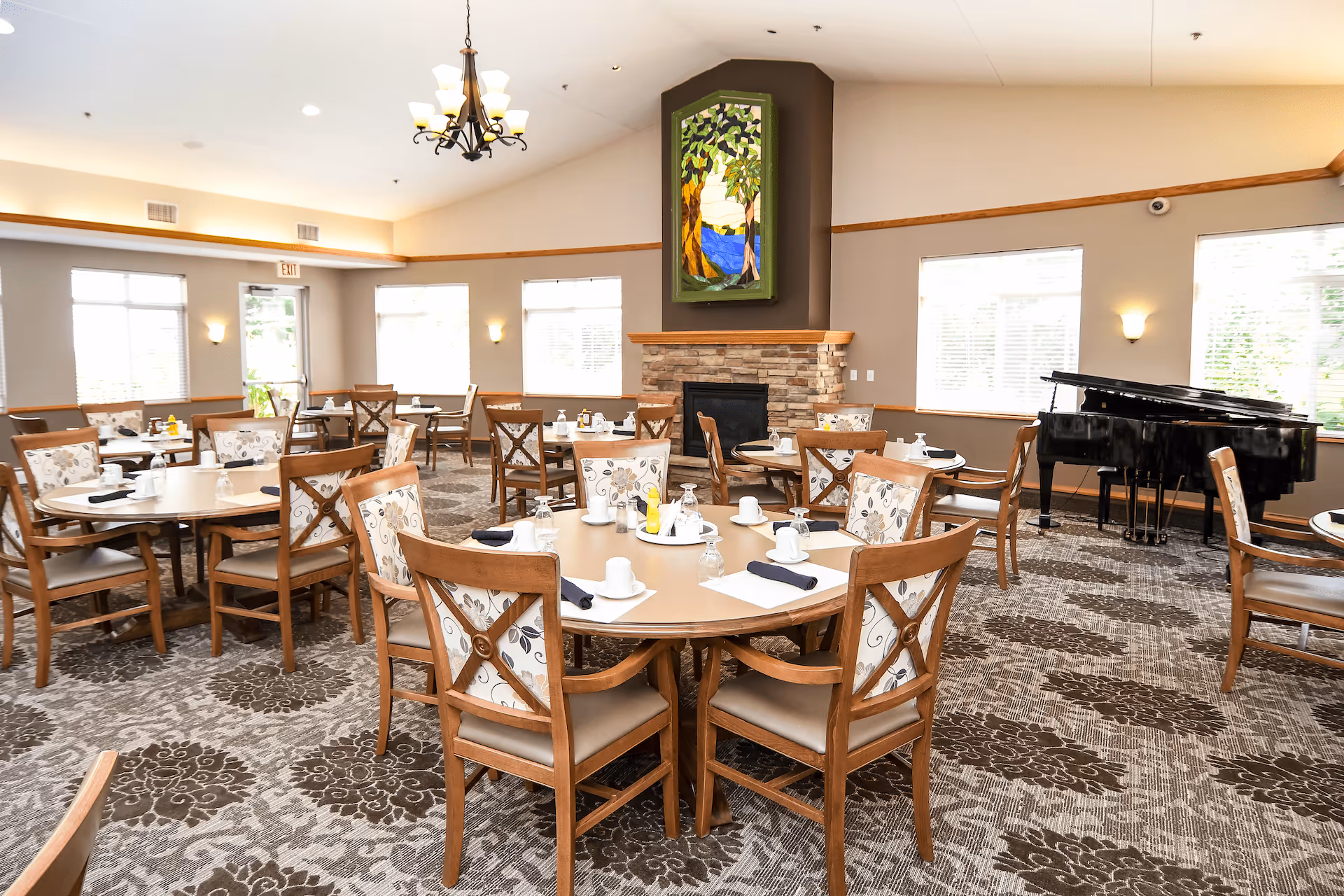 A bright and spacious dining room with multiple round tables set with white cups, napkins, and glassware. The room features wooden chairs with floral upholstery, a stone fireplace with a colorful stained glass artwork above it, large windows letting in natural light, and a black grand piano in the corner. The carpet has a floral pattern and a chandelier hangs from the ceiling.