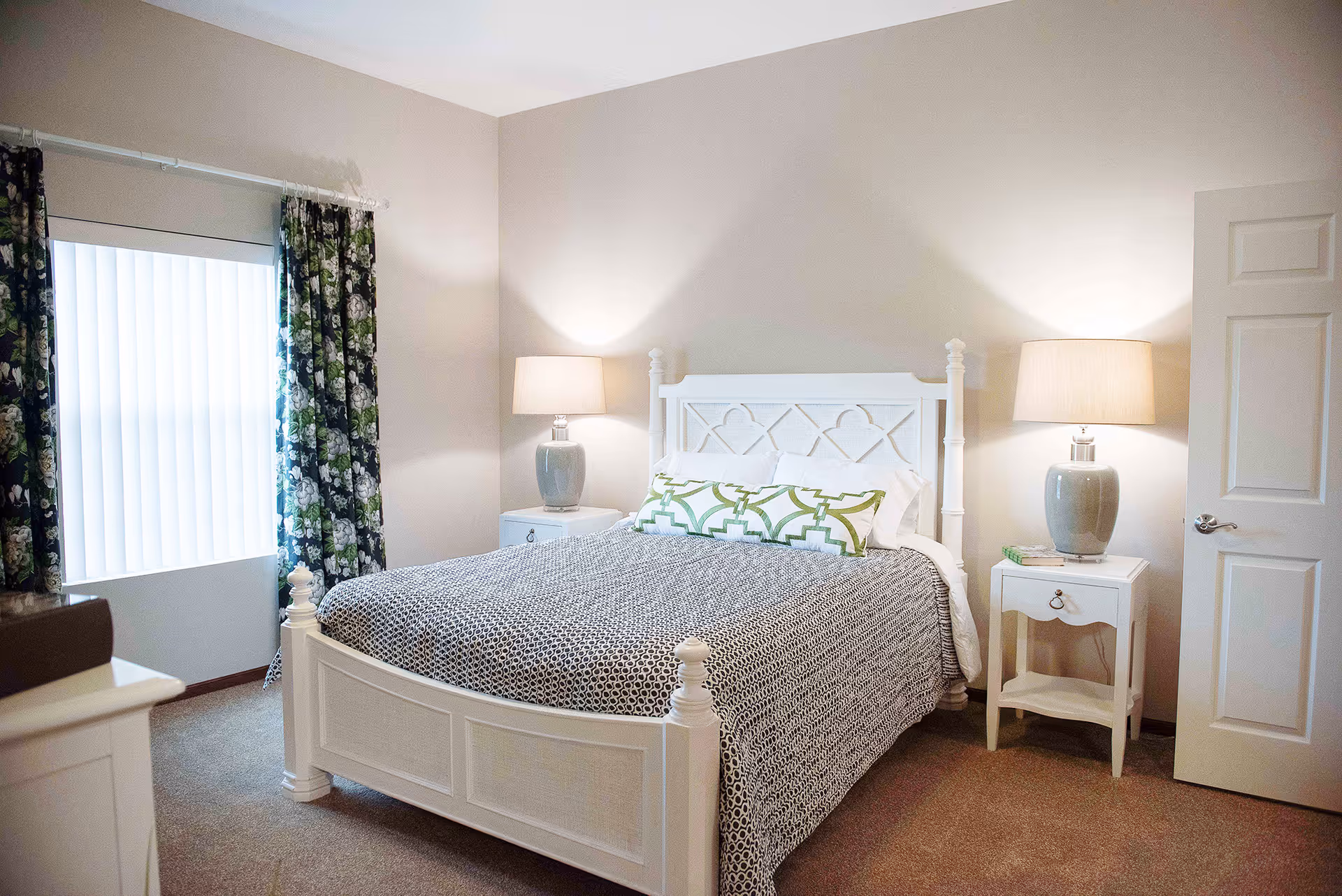 A neatly arranged bedroom with a white wooden bed frame, a patterned gray and white bedspread, and a long decorative pillow with a green geometric design. There are two matching white nightstands on either side of the bed, each with a gray lamp with a beige lampshade. A window with vertical blinds and floral curtains is visible on the left side, and a white door is partially open on the right side.