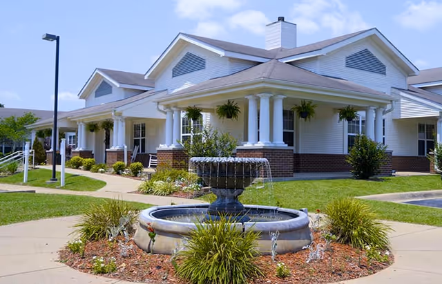 Exterior view of a single-story senior living facility building with white siding and brick accents. The building features a covered porch with white columns and hanging plants. In front of the building, there is a circular fountain surrounded by landscaped greenery and a paved walkway. The sky is clear and blue.