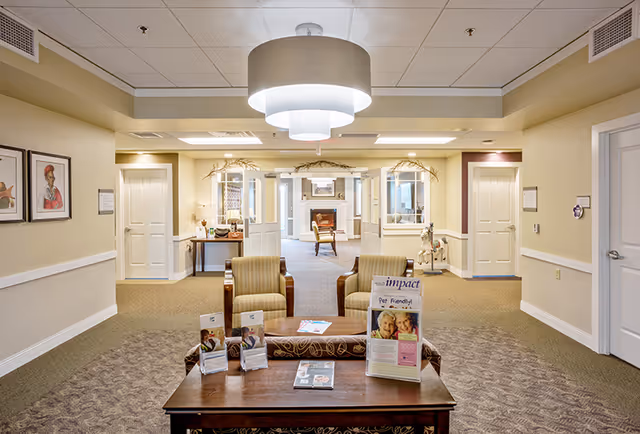 A senior living facility lobby with armchairs and a coffee table displaying brochures, leading to a hallway and additional seating.