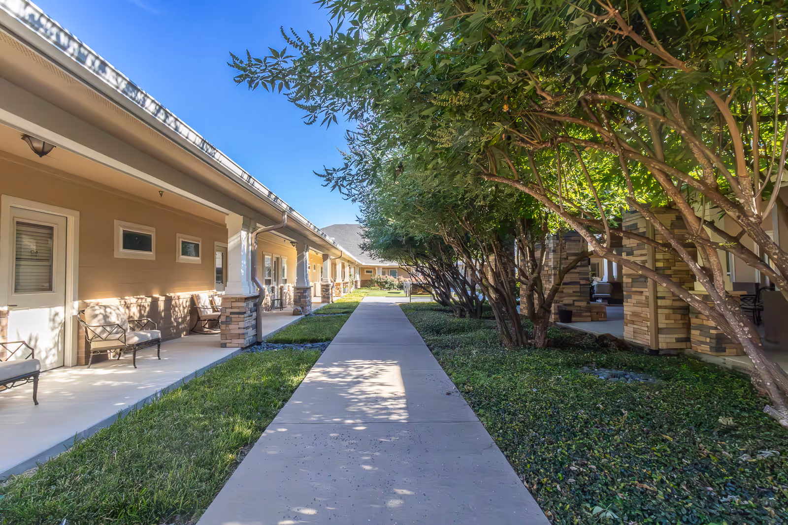 A paved walkway flanked by green grass and trees on the right side, with a row of beige buildings featuring covered patios and outdoor seating on the left side under a clear blue sky.