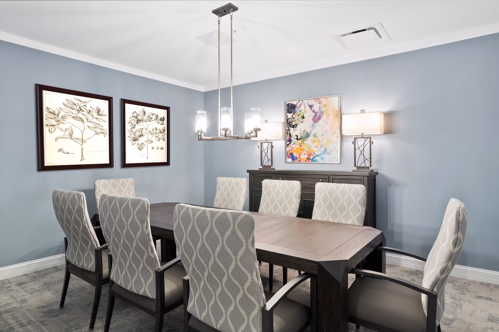 A modern dining room with a dark wooden rectangular table surrounded by eight upholstered chairs with patterned backs. The walls are painted light blue and decorated with three framed artworks. Two table lamps with rectangular shades sit on a dark wooden sideboard against the wall. A contemporary chandelier with six lights hangs above the table.