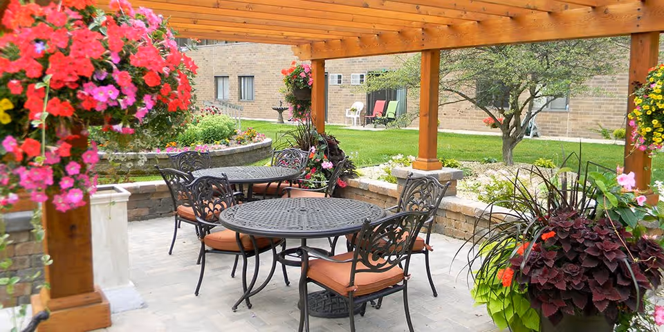 Outdoor patio area with metal tables and chairs with orange cushions under a wooden pergola. The space is surrounded by hanging flower baskets, potted plants, and a well-maintained garden with green grass and a tree. A brick building is visible in the background.