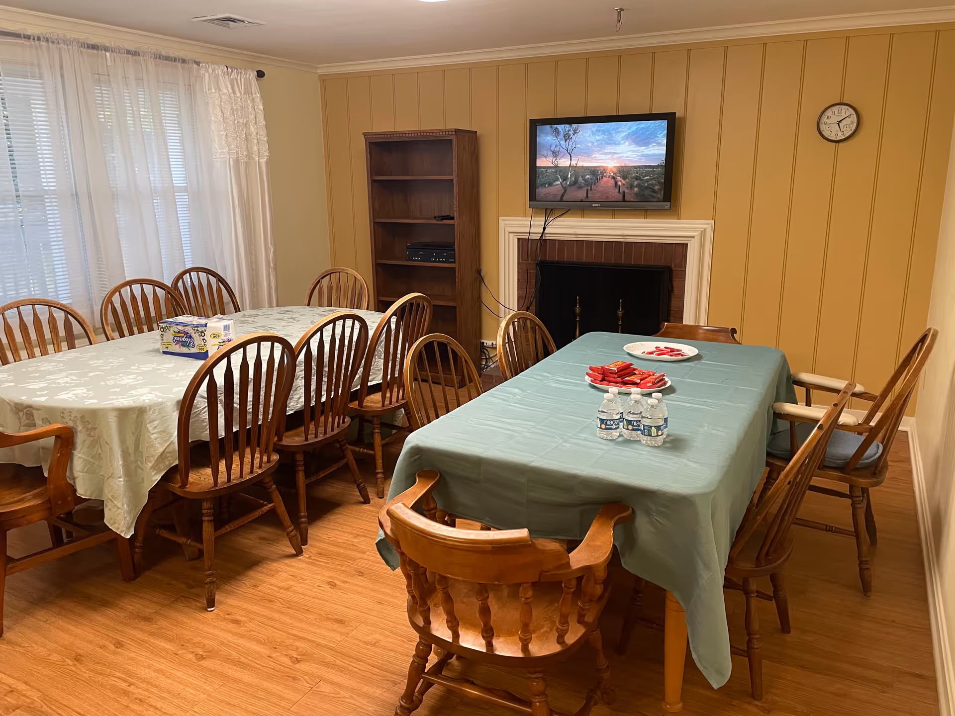 A dining room with two tables covered with tablecloths, surrounded by wooden chairs. One table has a white tablecloth with a box of tissues on it, and the other has a green tablecloth with bottled water and plates of snacks. There is a wooden bookshelf and a wall-mounted TV above a fireplace on the far wall. A clock is mounted on the wall to the right.