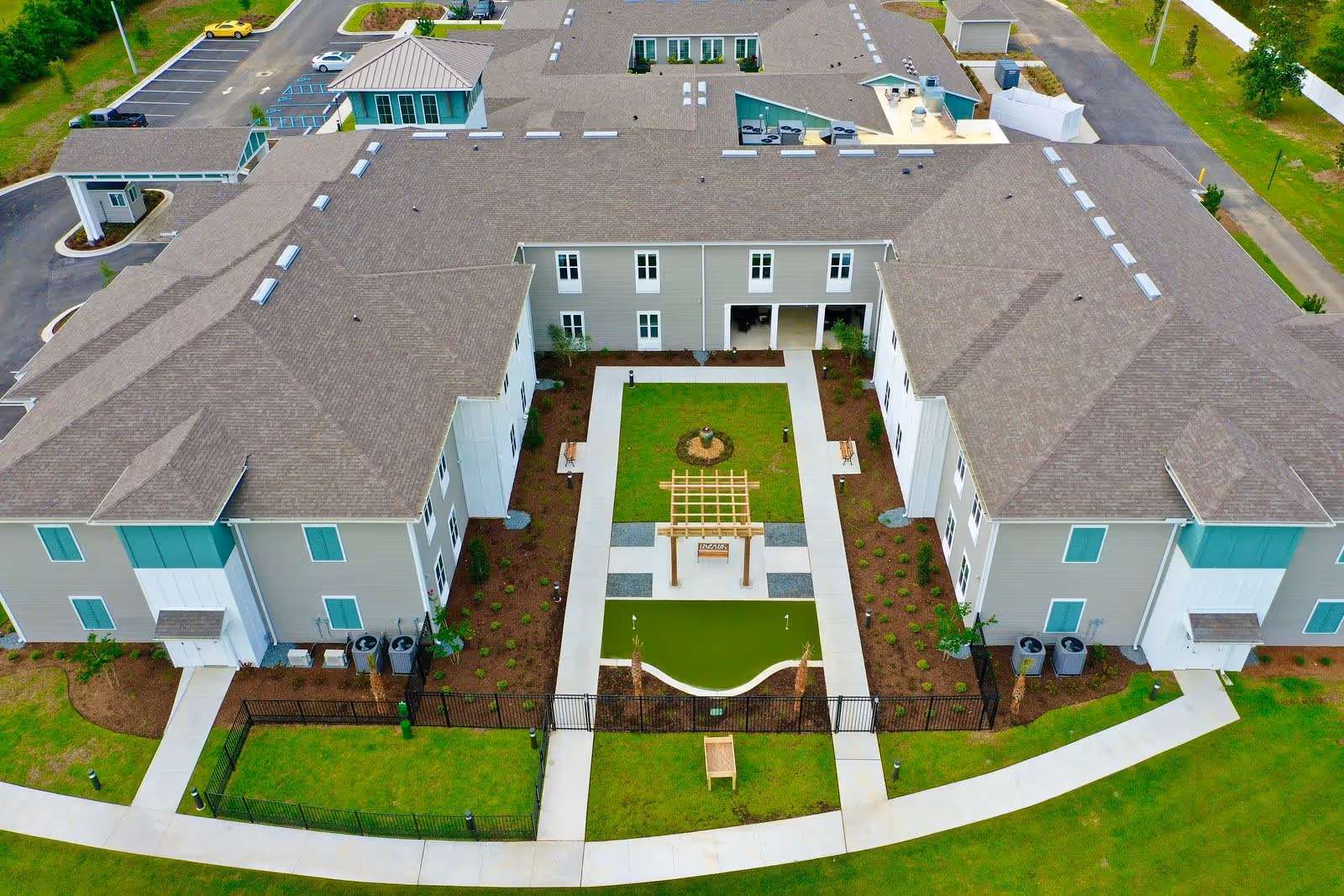 Aerial view of The Canopy at Azalea Grove senior living facility showing a large U-shaped building surrounding a landscaped courtyard with a small putting green, benches, and a wooden pergola. The building has multiple windows and a gray roof, with parking spaces visible in the background.