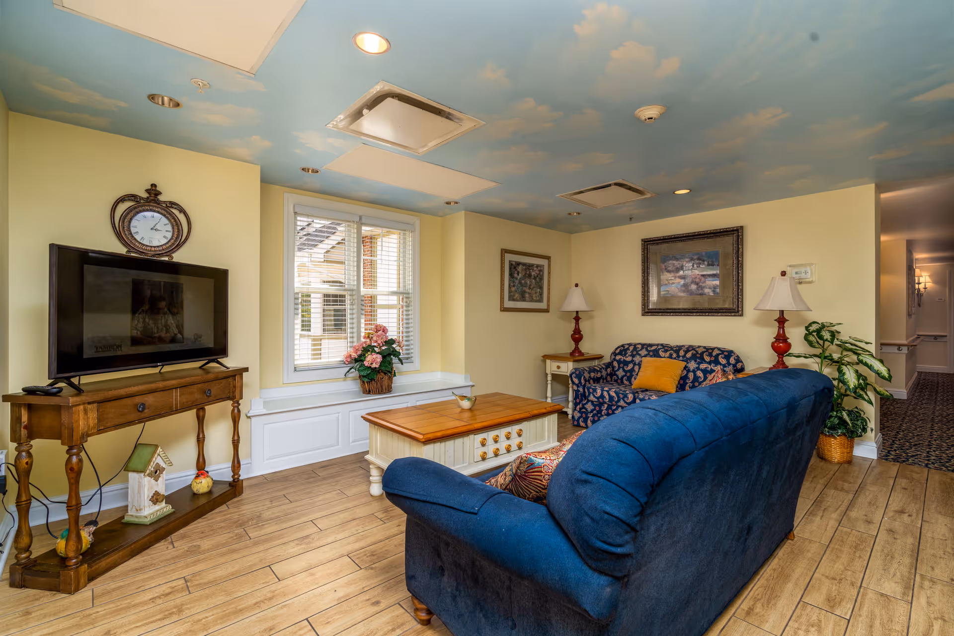 A cozy living room with a blue couch and a patterned loveseat, a wooden coffee table, two side tables with red lamps, a flat-screen TV on a wooden stand, a window with white blinds, framed artwork on the walls, a potted plant, and a ceiling painted with a sky and clouds design.