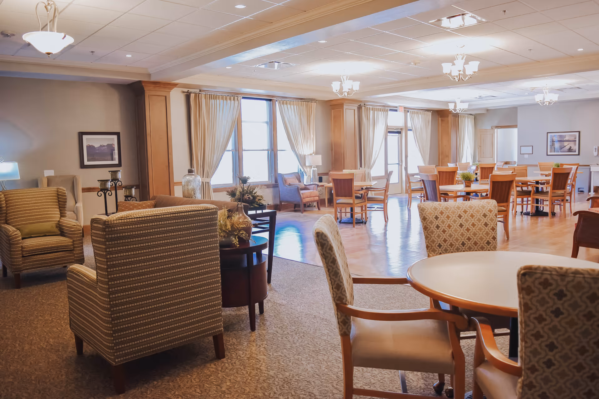 A spacious senior living common area with a mix of seating options including patterned armchairs, wooden chairs, and round tables. The room features large windows with beige curtains allowing natural light to fill the space, wooden flooring in the dining area, and carpet in the lounge area. The walls are decorated with framed artwork and the ceiling has multiple light fixtures.