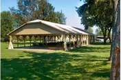 Open-sided pavilion with a sloped roof and picnic tables sits on a grassy lawn.