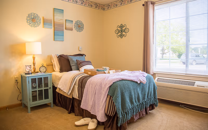 A neatly made single bedroom with a bed, bedside table and lamp beside a large window.