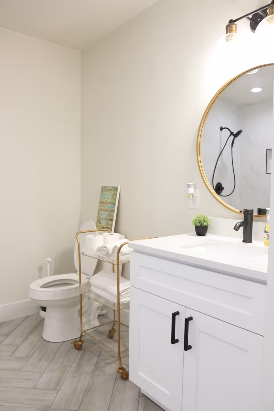 A clean and modern bathroom featuring a white toilet, a gold metal cart with towels and toilet paper, a white vanity with black handles, a round mirror with a gold frame, and a black faucet. The floor has light gray tiles arranged in a herringbone pattern.