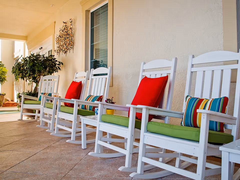 White wooden rocking chairs with green seat cushions and colorful pillows lined up on a covered porch.