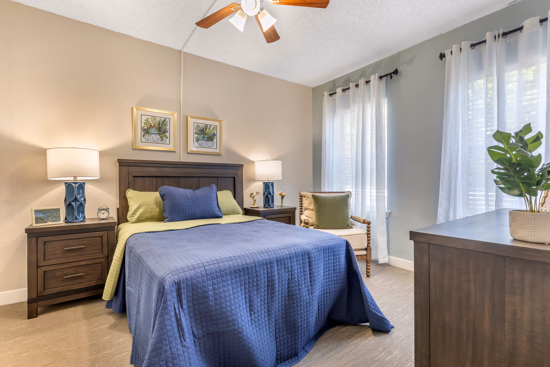 Sunlit bedroom with a blue-covered bed, wooden nightstands and lamps, a chair by sheer-curtained windows, and a dresser with a plant.