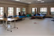 A spacious physical therapy or rehabilitation room with large windows along two walls allowing natural light. The room contains therapy tables, exercise balls, and various rehabilitation equipment on a light-colored floor. The walls are beige with a decorative border near the ceiling.