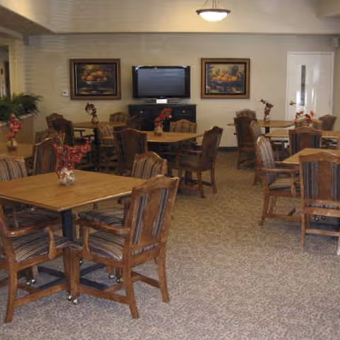 A dining room with multiple wooden tables and cushioned chairs arranged neatly. Each table has a small floral centerpiece. The room has carpeted flooring, two framed paintings on the wall, a flat-screen TV mounted on a cabinet, and a ceiling light fixture.