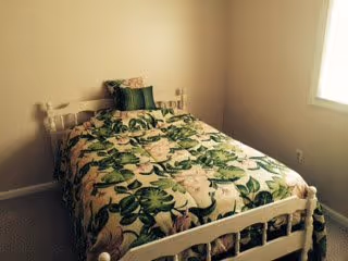 A small bedroom with a single white wooden bed frame and a floral patterned bedspread featuring green leaves and light pink flowers. There is a matching pillow on the bed. The walls are plain and light-colored, and there is a window on the right side letting in natural light.