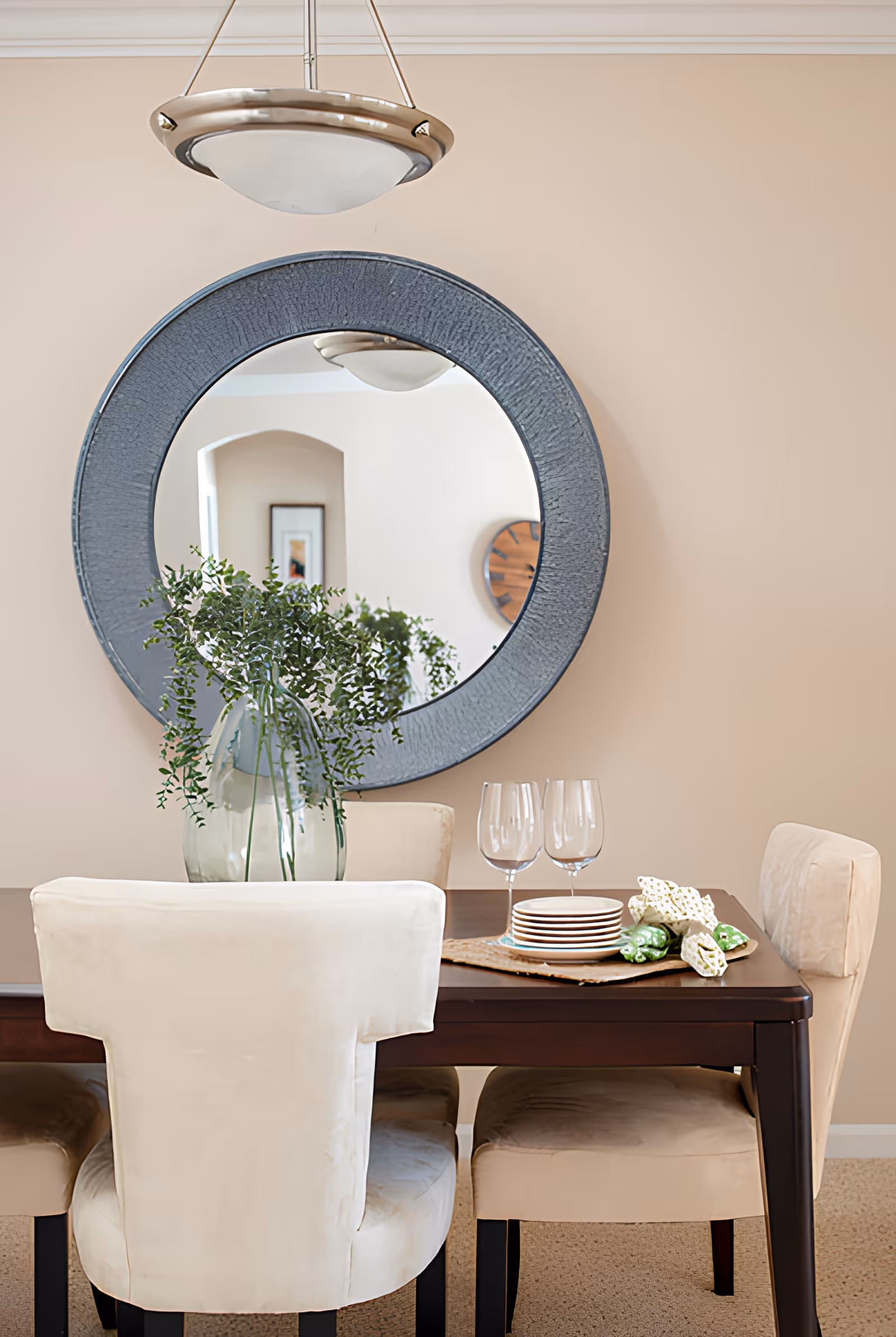 A dining room with a dark wooden table and four beige upholstered chairs. On the table, there is a glass vase with green leafy branches, two empty wine glasses, a stack of small plates, and some decorative items. A large round mirror with a textured blue-gray frame hangs on the beige wall behind the table, reflecting part of the room including a wall clock and a framed picture.