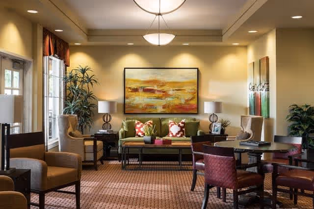 A well-lit living room area with a green sofa adorned with red and white patterned pillows, two beige armchairs, and a coffee table with decorative plants. The walls are painted light yellow and feature two large abstract paintings. There are also round tables with red chairs, potted plants, and two table lamps with white shades. Large windows allow natural light to enter the room.