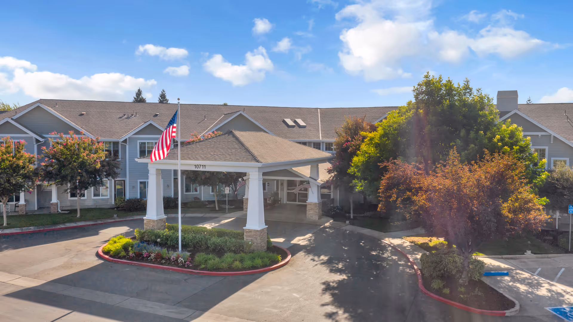 Exterior view of The Commons on Thornton senior living facility showing a two-story building with a covered entrance, an American flag on a flagpole, landscaped greenery, and a clear blue sky with some clouds.