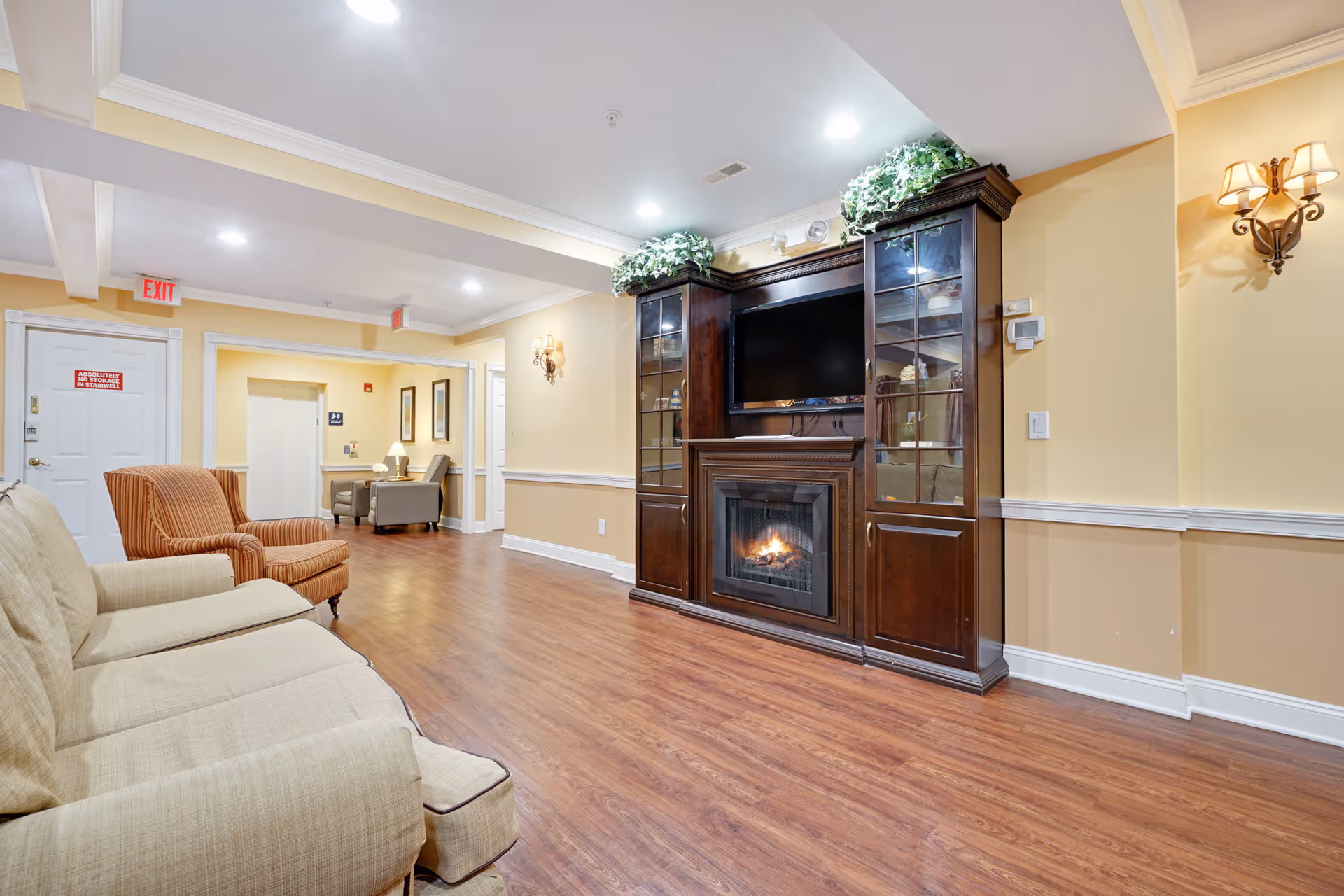 A cozy living room area in Fox Trail Memory Care Living featuring a beige sofa and an orange armchair on wooden flooring. A large dark wood entertainment center with a built-in fireplace and a flat-screen TV is against the wall, decorated with green plants on top. The walls are painted light yellow with white trim, and there are wall sconces providing warm lighting. In the background, there is a small seating area with two gray chairs and a table with a lamp.