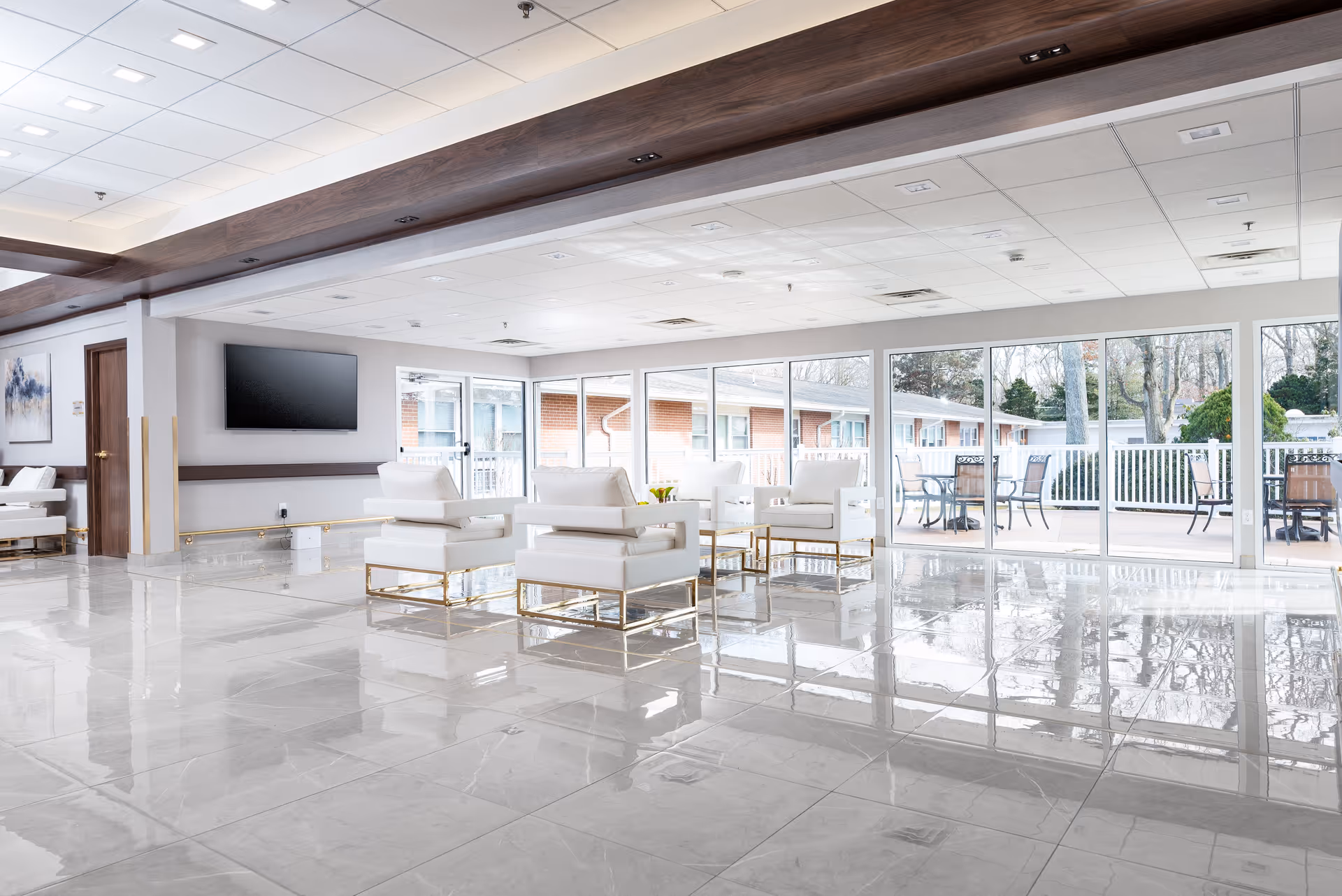 A bright and spacious living room area with white modern chairs arranged around a glass coffee table. Large windows and sliding glass doors provide a view of an outdoor patio with tables and chairs. The room has a glossy tiled floor, a mounted flat-screen TV on the wall, and a ceiling with recessed lighting.